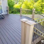 A large, curved backyard deck built with DIY deck materials features brown composite flooring and beige railings overlooking a green lawn and trees. A black metal chair sits in the corner near the house.