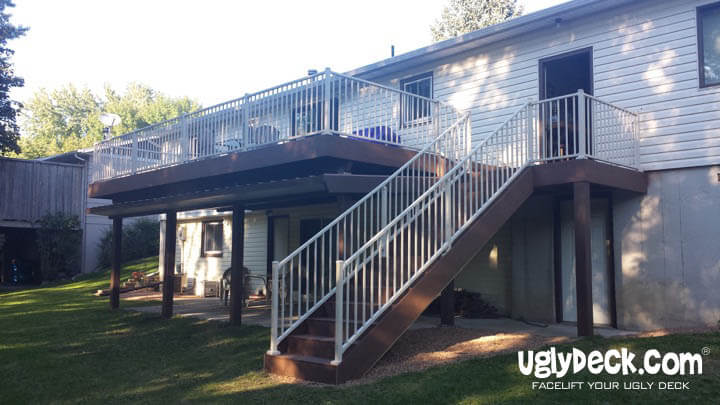 A two-story house with a large elevated deck, professionally crafted by an expert Deck Builder. The deck features brown flooring, white railings, and a staircase leading down to the grassy backyard, with a shaded area below.