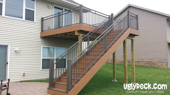 A raised backyard deck with brown flooring and gray railings, featuring a staircase leading to the grassy yard. The deck is attached to a light-colored house. Text reads: uglydeck.com, FACELIFT YOUR UGLY DECK with premium DIY Deck Materials.