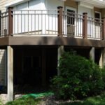 A two-story house with a brown raised deck, built by expert deck builders, features metal railings, stairs to the yard, and green bushes beneath. The UglyDeck.com logo appears in the lower right corner.