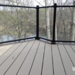 View from a deck with light-colored, wood-like flooring and black railings, overlooking leafless trees and a calm body of water. Perfect inspiration for your DIY deck materials project. The uglydeck.com logo is at the bottom right corner.