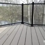 View from a deck with light-colored, wood-like flooring and black railings, overlooking leafless trees and a calm body of water. Perfect inspiration for your DIY deck materials project. The uglydeck.com logo is at the bottom right corner.