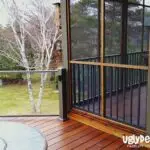 A wooden deck with glass and black railings overlooks a grassy yard with trees and a lake in the background. The partially covered deck features a table—perfect for DIY Deck Materials or your next Deck Install. UglyDeck.com logo is in the bottom right corner.
