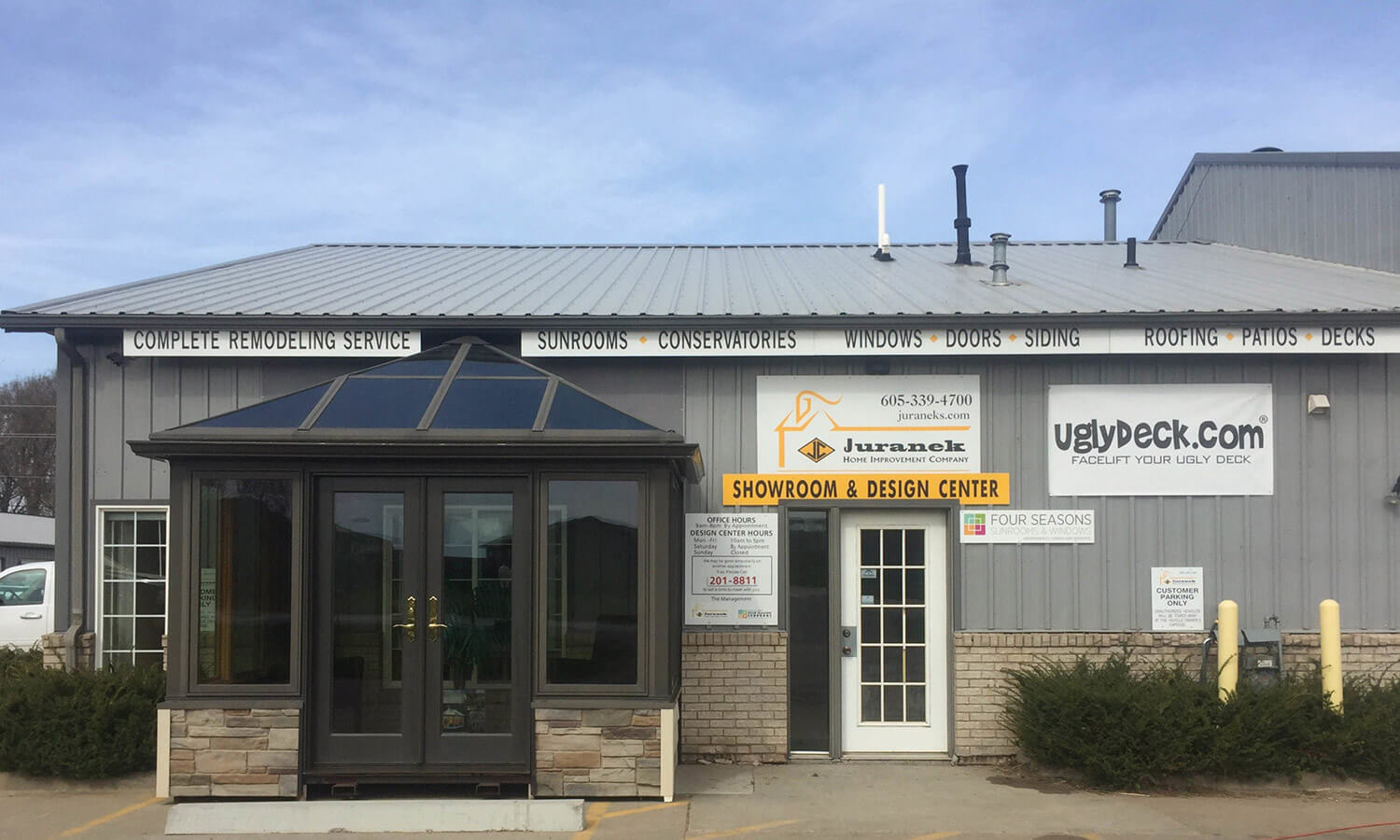 A gray commercial building with a glass entryway, signs for home remodeling and deck install services, and a banner reading Complete Remodeling Service above the windows. Shrubs line the front of the building.