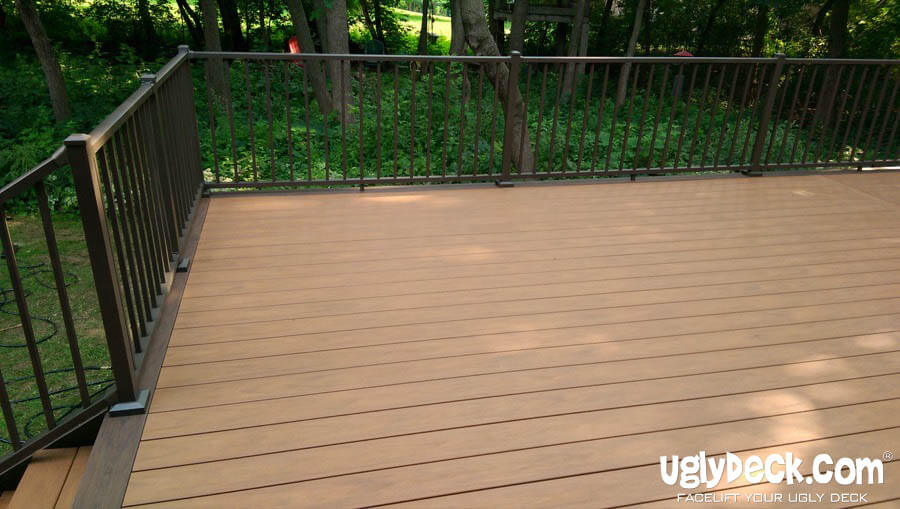 A spacious wooden deck with dark metal railings overlooks a green, wooded backyard. Perfect for DIY Deck Materials or a professional Deck Install, the clean deck glows in sunlight. UglyDeck.com branding appears in the lower right corner.