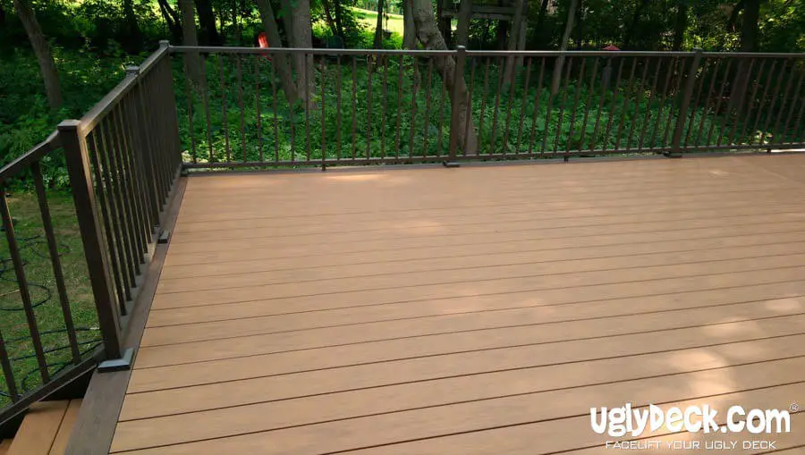 A spacious wooden deck with dark metal railings overlooks a green, wooded backyard. Perfect for DIY Deck Materials or a professional Deck Install, the clean deck glows in sunlight. UglyDeck.com branding appears in the lower right corner.