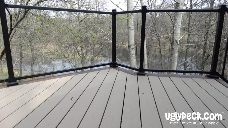 A deck with beige composite boards and a black-framed glass railing, built using quality DIY Deck Materials, overlooks trees and a small body of water. The words uglydeck.com appear in the lower right corner.