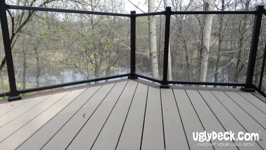 A deck with beige composite boards and a black-framed glass railing, built using quality DIY Deck Materials, overlooks trees and a small body of water. The words uglydeck.com appear in the lower right corner.
