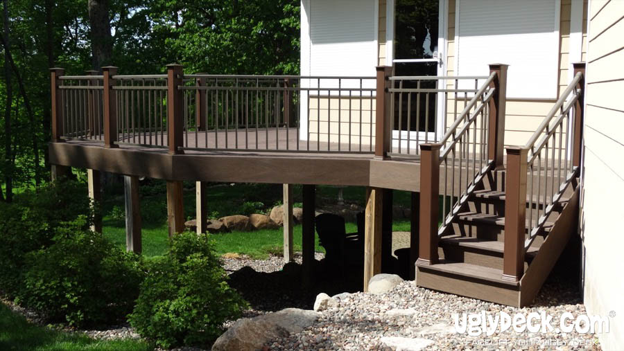 A raised wooden deck with brown railings and steps, attached to a beige house. Perfect for your next DIY Deck Materials project, it overlooks a landscaped yard with rocks, shrubs, and trees. The logo uglydeck.com appears in the bottom right corner.