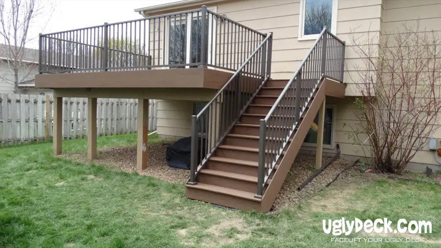 A raised wooden deck with metal railings and a staircase leads to the backyard of a beige house. Built by a professional Deck Builder, the deck is supported by posts, with rocks underneath and grass surrounding the area. uglydeck.com is visible in the corner.