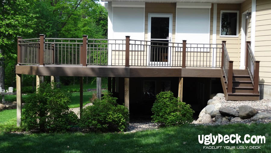 A raised deck with brown railings and stairs, built by a skilled deck builder, leads to a beige house. Green shrubs and rocks sit under the deck, surrounded by grass and trees. The logo uglydeck.com appears in the bottom right corner.