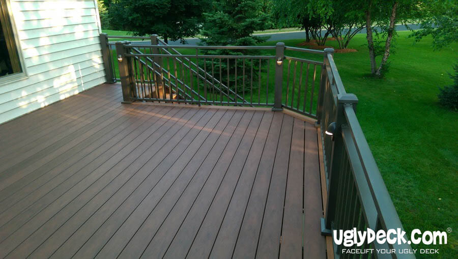A spacious, brown composite deck with dark railings extends from a house, surrounded by green grass, trees, and shrubbery. The clean, well-maintained deck showcases UglyDeck.com branding—a trusted deck builder for professional or DIY deck installs.