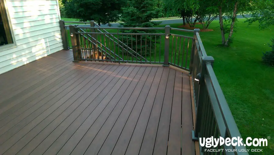 A spacious, brown composite deck with dark railings extends from a house, surrounded by green grass, trees, and shrubbery. The clean, well-maintained deck showcases UglyDeck.com branding—a trusted deck builder for professional or DIY deck installs.