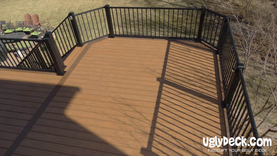 A brown composite deck with black metal railings, installed by a skilled Deck Builder, overlooks a grassy yard with outdoor furniture in the distance. The deck is clean and spacious. uglydeck.com is visible in the lower right corner.