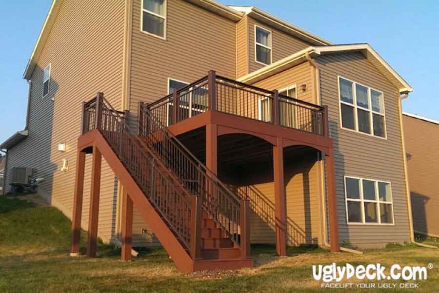A two-story house with tan siding features a raised wooden deck by a professional deck builder, complete with dark railings and a staircase to the backyard. The shaded space beneath enhances outdoor living, and the UglyDeck.com logo appears in the corner.