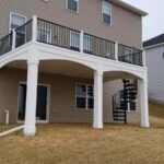 A beige two-story house features a white-railed elevated deck built with premium DIY Deck Materials, supported by two columns and a black spiral staircase that leads to the grassy backyard on a sloped lot.