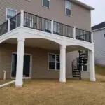 A beige two-story house features a white-railed elevated deck built with premium DIY Deck Materials, supported by two columns and a black spiral staircase that leads to the grassy backyard on a sloped lot.