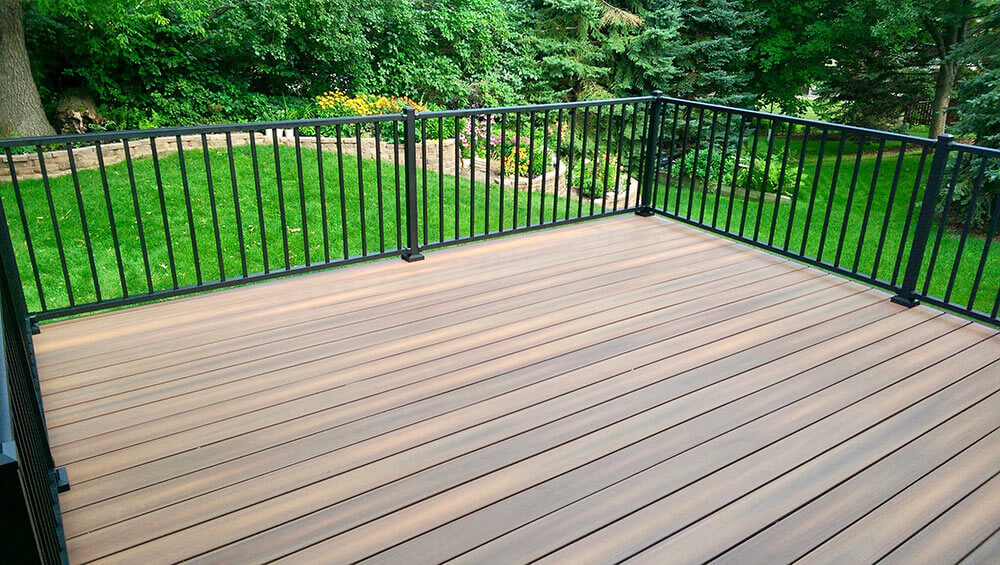 A spacious outdoor deck with wood-look flooring and black metal railings overlooks a lush green lawn. Perfect for those considering DIY deck materials or planning a new deck install to enhance their landscaped garden with trees and shrubs.