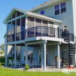A two-story house features a screened-in upper deck, built with quality DIY deck materials, black spiral staircase, and patio furniture below. The yard is beautifully landscaped with grass, shrubs, and a small rock garden.