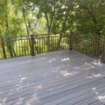 A gray composite deck built by expert deck builders with metal railings overlooks lush green trees; sunlight filters through the foliage, creating a peaceful outdoor space. The bottom right corner features the uglydeck.com logo.
