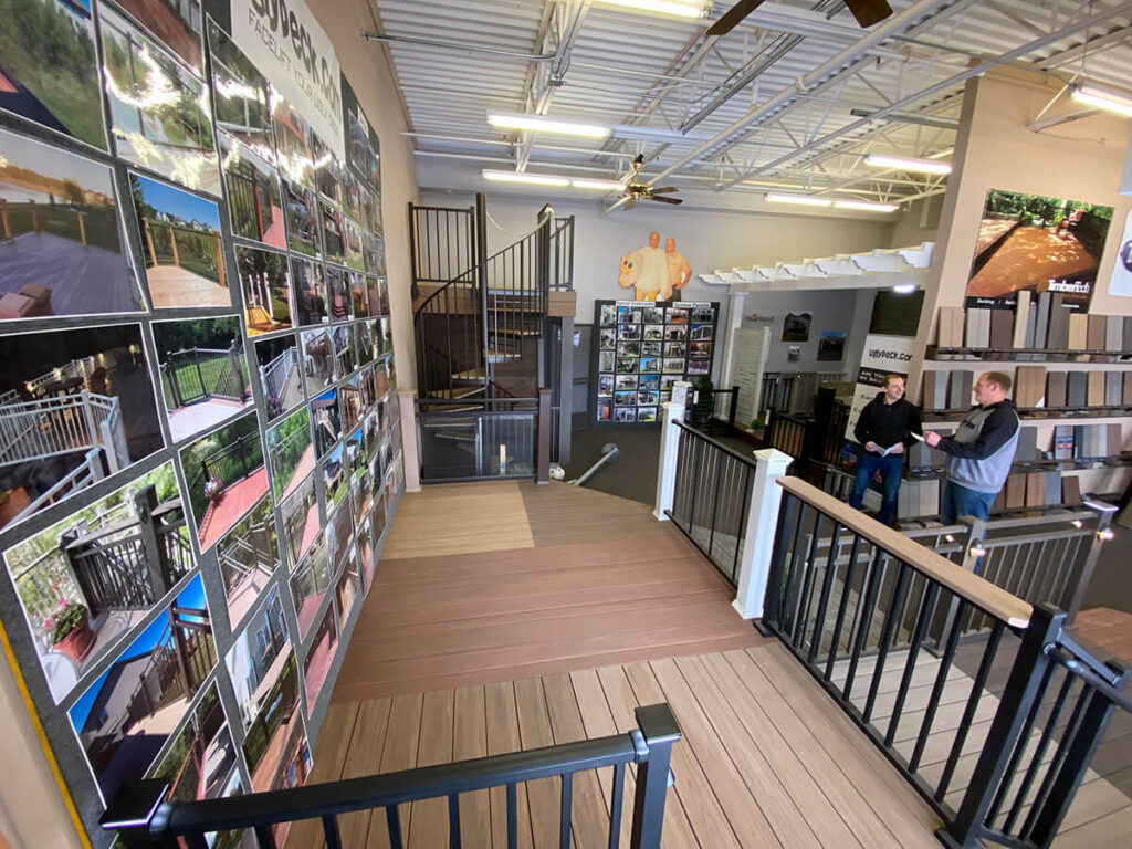 A showroom displays deck railings and flooring samples for DIY deck materials. Two people stand near a counter, while walls are lined with photos of completed decking projects. Ceiling fans and bright overhead lights illuminate the space.