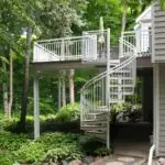 A modern two-story deck with white railings and a spiral staircase overlooks a lush garden. Expert deck builder craftsmanship ensures the lower level is shaded and partially covered by the upper deck, blending style and function.