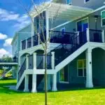 A modern two-story house with gray siding, white trim, and a large elevated deck built by a professional deck builder. A small, leafless tree stands in the green lawn under a blue sky with clouds.