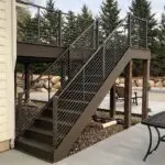 A modern outdoor staircase with dark brown steps and metal cable railing leads to an elevated deck. Built by a skilled Deck Builder, the deck is supported by wooden posts, with trees and patio furniture in the background.