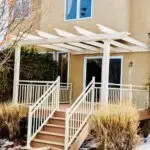 A beige pergola with white railings covers a raised deck, expertly crafted by a local Deck Builder, with stairs leading to a sliding glass door on a tan house. Bushes and trees frame the deck, and a light dusting of snow coats the ground.