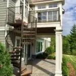 A two-story house with gray siding features a brown metal spiral staircase leading from a covered patio to an upper balcony, perfect for a future Deck Install. Landscaping includes green shrubs, small trees, and stone pavers.