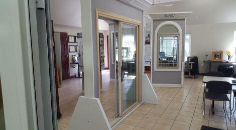A showroom displays various window and door models, including a large sliding glass door perfect for any Deck Builder or DIY Deck Materials project, with office furniture and tiled floors visible in the background.