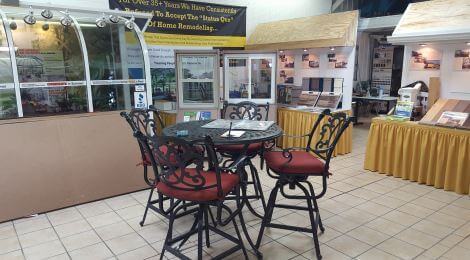 A showroom displays home renovation products, including windows, doors, siding, and DIY deck materials. In the center is a round glass-top table with four ornate metal chairs with red cushions, set on a tiled floor.