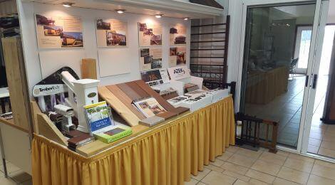 A display table covered with a yellow skirt showcases DIY Deck Materials and brochures, along with informational boards for deck builders and deck install advice, set in a showroom with tiled floors and glass doors.