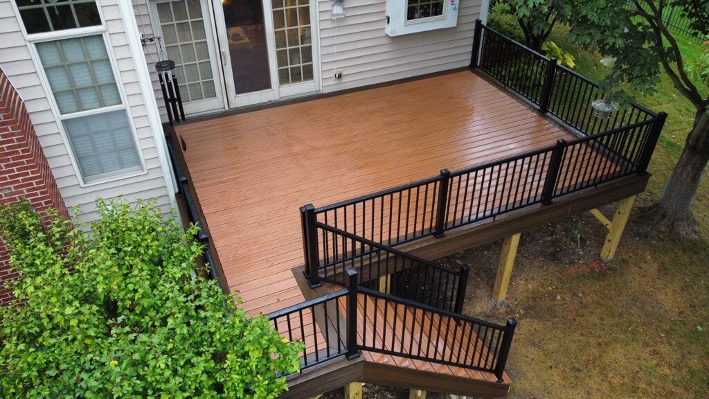 A raised wooden deck with black railings, expertly crafted by a professional deck builder, is attached to a beige house. Stairs lead down to the yard, and the deck is surrounded by lush green trees and grass.