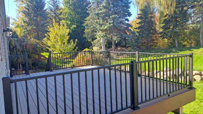 A spacious, wooden backyard deck with black railings overlooks a green lawn and trees on a sunny day—perfect inspiration if you’re considering a professional deck builder or quality DIY deck materials for your own outdoor retreat.