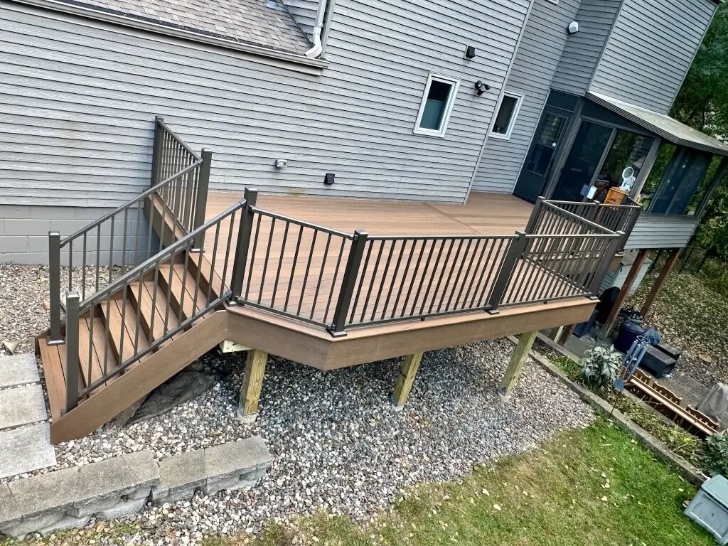 A raised wooden deck with metal railings, expertly crafted by a Deck Builder, is attached to a gray house. Stairs lead down to a rocky area, and the deck is supported by wooden posts. A screened porch sits to the right above a grassy yard.