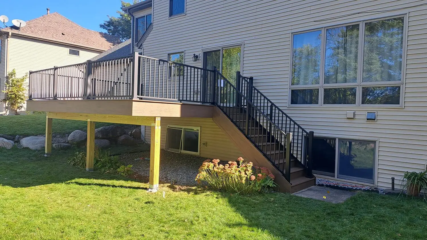 A raised wooden deck install with black metal railings and stairs leads to a grassy backyard, attached to a beige house with large windows and garden plants below the deck—perfect inspiration for your next DIY deck materials project.