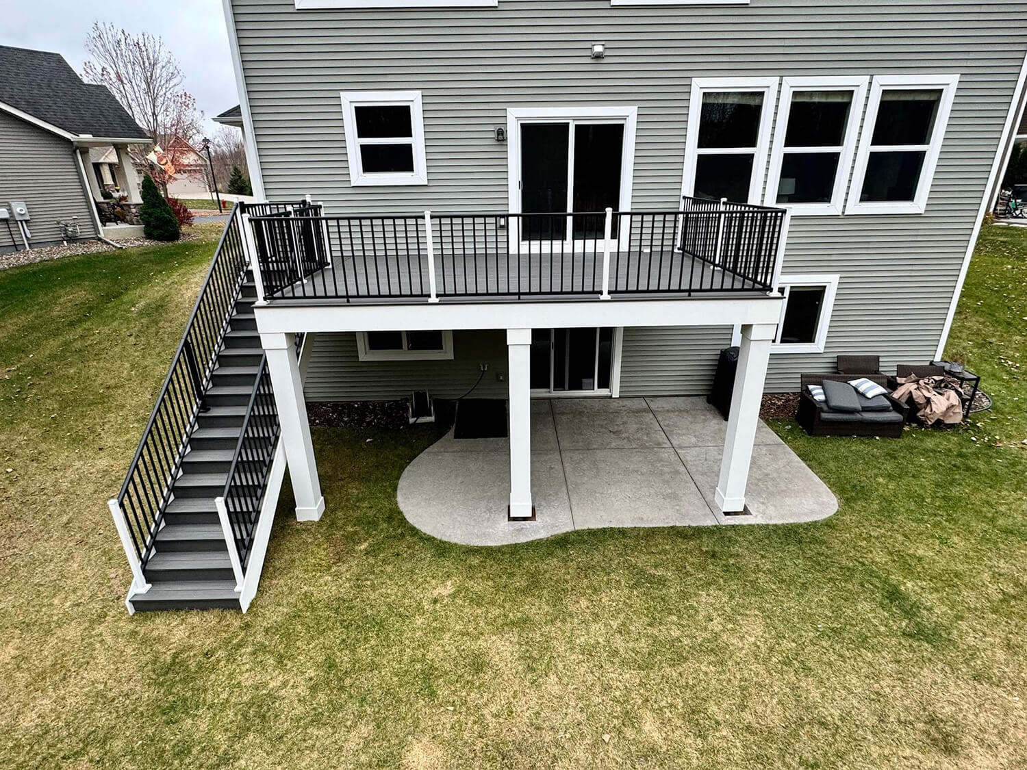 A two-story house with a raised deck install featuring black railings and stairs leading to the grassy, sloped backyard. Under the deck is a concrete patio with a seating area on the right side—perfect for enjoying your outdoor space.