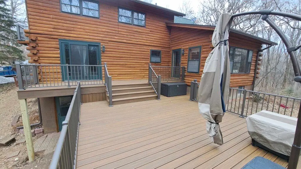A spacious wooden deck, built with quality DIY Deck Materials, extends from a log cabin-style house. Stairs lead to a door, and covered patio furniture and leafless trees are visible in the background.