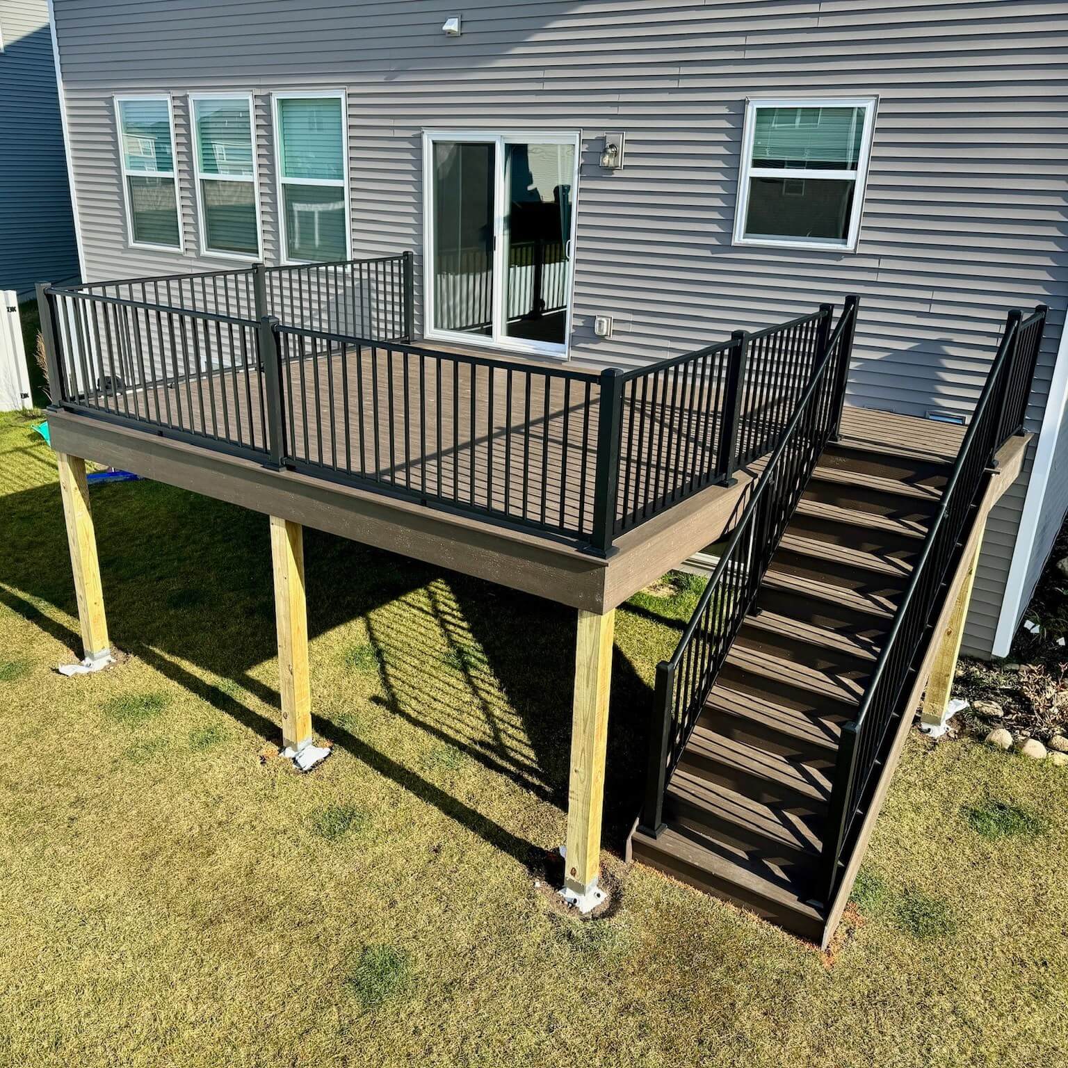 A raised wooden deck installed by a professional deck builder, featuring black metal railings and a staircase leading to a grassy backyard. The deck is supported by four wooden posts anchored to concrete footings.