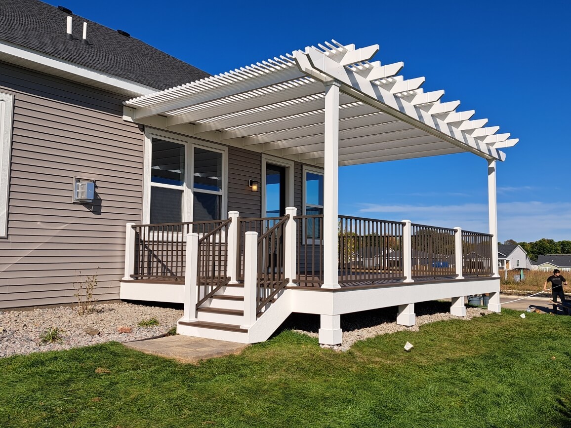 A modern house with a raised wooden deck, expertly crafted by a professional deck builder, featuring white railings and a white pergola, accessed by stairs, surrounded by green grass and under a clear blue sky.