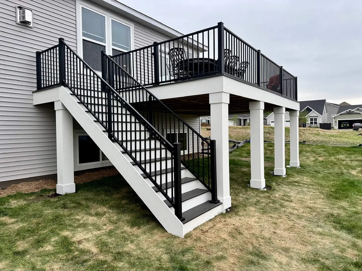 A raised backyard deck with black railings, white posts, and stairs leading down to a grassy yard. Built with quality DIY Deck Materials, the deck features outdoor furniture and is attached to a gray house with large windows.