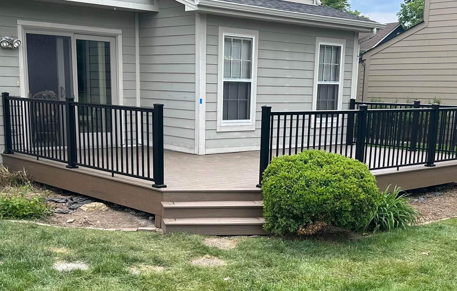 A backyard deck install features black metal railings, brown wooden flooring, and steps to a grassy yard. Attached to a light gray house with large windows and sliding glass doors, the deck is bordered by shrubs and plants.