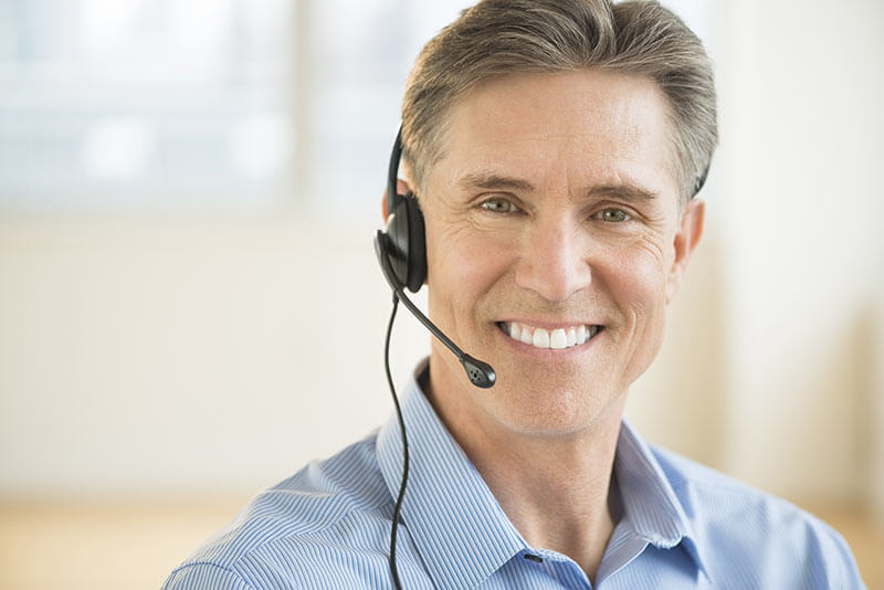 Smiling man wearing a headset with a microphone, dressed in a light blue shirt, looking at the camera in a bright, blurred office setting—ready to assist with deck install questions or recommend quality DIY deck materials.