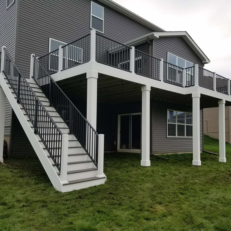 A two-story house with gray siding features a raised white deck built with quality DIY Deck Materials, black railings, a staircase leading to a grassy yard, and an open area beneath the deck with sliding glass doors.