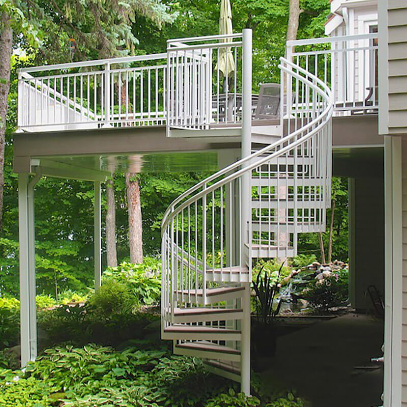 A white metal spiral staircase, built by a skilled deck builder, leads from the patio beneath a raised deck to the upper deck, surrounded by lush green trees and plants in a peaceful backyard setting.