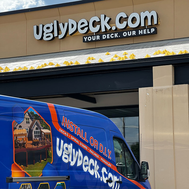 A blue UglyDeck.com van, showcasing deck builder services and DIY deck materials, is parked in front of a building with a large sign reading "YOUR DECK. OUR HELP." The van displays images of decks and advertises installation and DIY services.