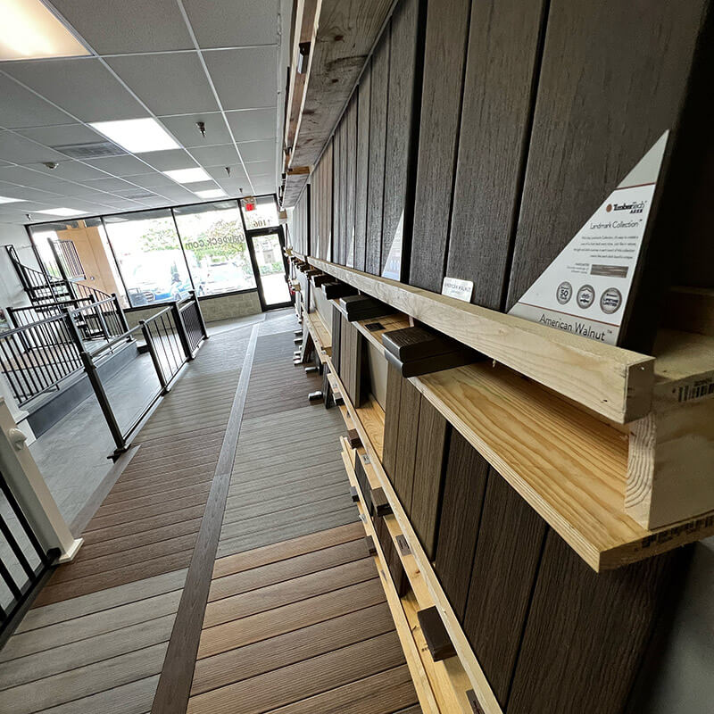 A display wall in a showroom exhibits samples of composite decking boards in various colors and wood textures—perfect for deck builders or those seeking DIY deck materials—with natural light streaming through the glass entrance door.