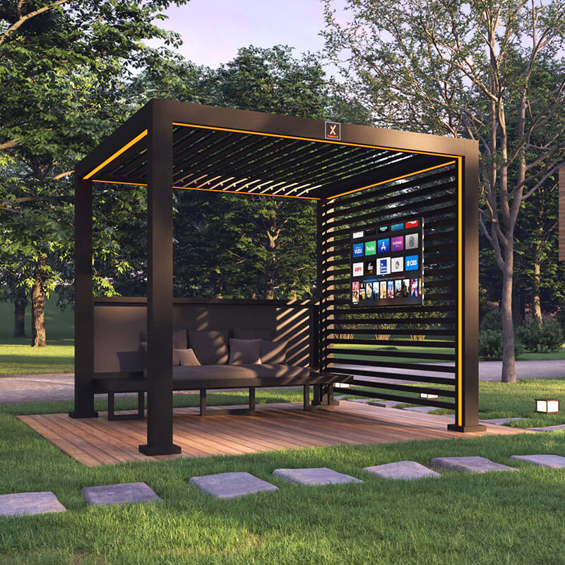 A modern outdoor gazebo with a slatted roof and walls sits on a wooden platform crafted by expert deck builders. Inside, there's a black sofa and a large flat-screen TV. The area is surrounded by grass and trees.
