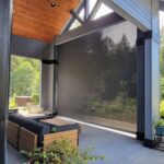 A modern covered patio with a large retractable mesh screen, outdoor seating, a coffee table, and views of trees and greenery. Sunlight filters through the screen and pergola, creating welcome shade for this relaxing space.